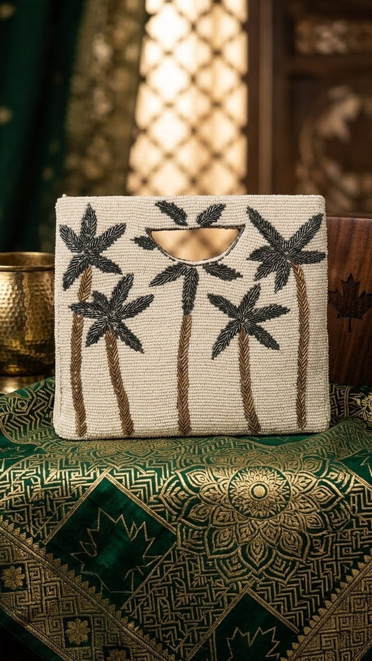 Oh my Palm Beaded Bag — A rectangular beige clutch bag with black palm tree embroidery and brown trims, resting on a green and gold patterned textile with a warm, bohemian background. — Zohricanada