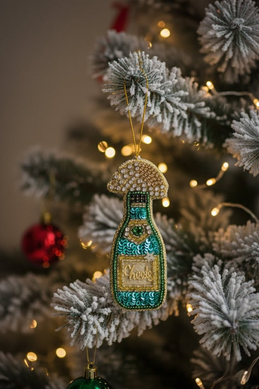 Champagne Bottle Ornament — A decorative Christmas ornament shaped like a bottle with a mushroom cap, hanging on a flocked Christmas tree with warm string lights. The ornament features turquoise, gold, and white beaded details. — Zohricanada