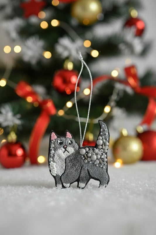 Gray Cat Ornament — A gray and white cat-shaped Christmas ornament with glittery details hangs from a silver string in front of a blurred decorated Christmas tree with red and gold ornaments. — Zohricanada
