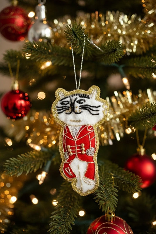 Nutcracker Cat Ornament — A festive embroidered cat ornament dressed in a red military-style uniform with gold trim, hanging on a decorated Christmas tree with lights and red ornaments in the background. — Zohricanada