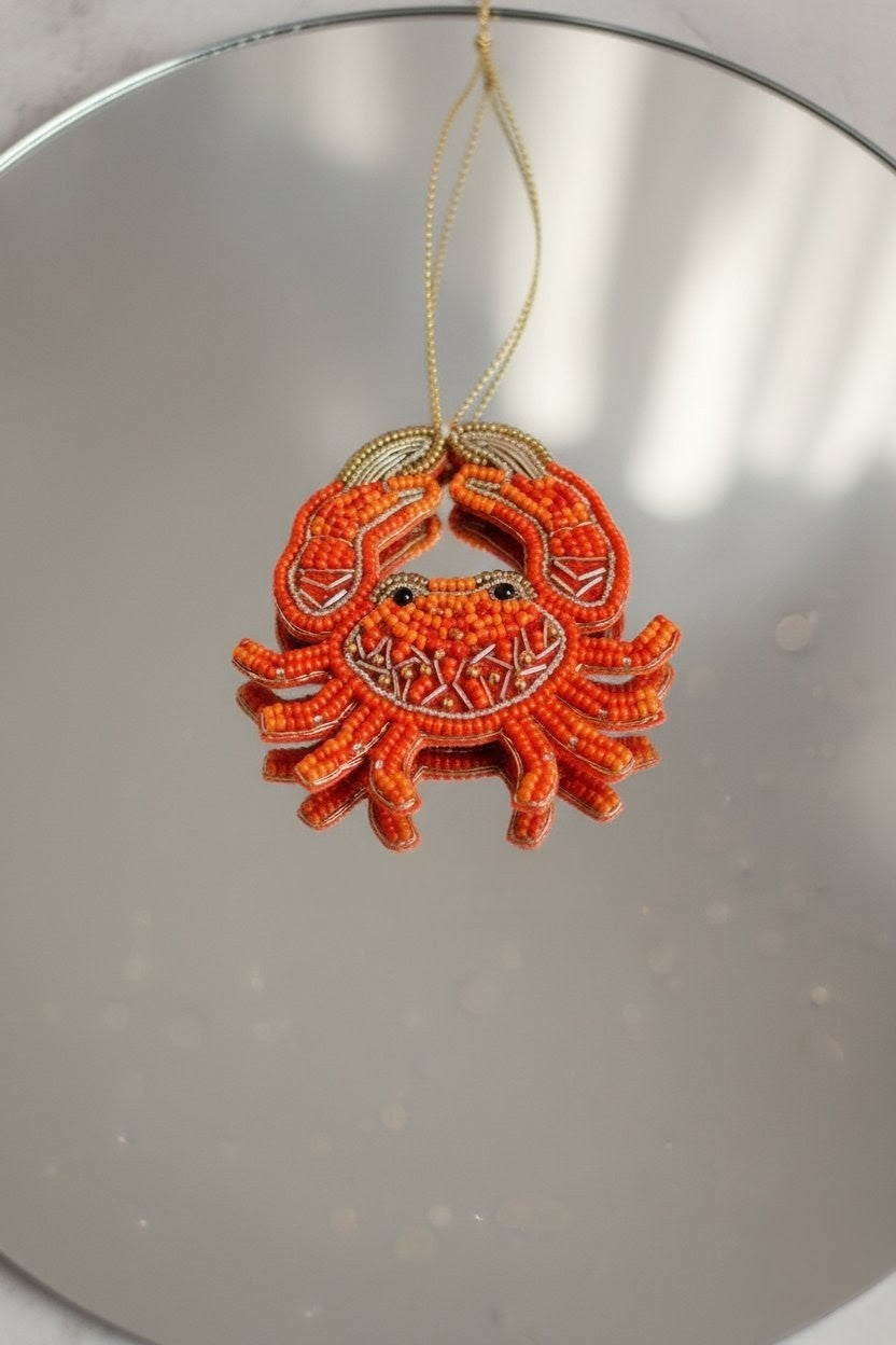Orange Crab Ornament — A small orange beaded crab ornament with a gold hanging string, photographed against a reflective silver surface. — Zohricanada