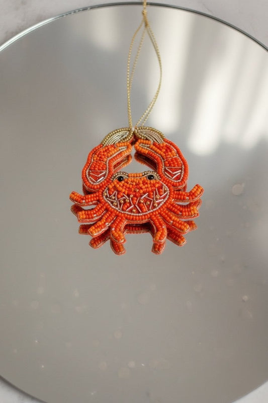 Orange Crab Ornament — A small orange beaded crab ornament with a gold hanging string, photographed against a reflective silver surface. — Zohricanada