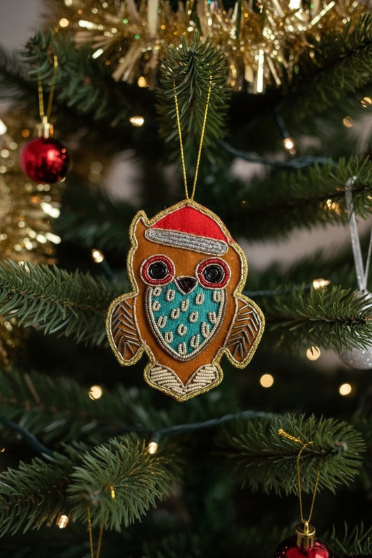 Owl Ornament — A handmade embroidered owl Christmas ornament hanging on a decorated Christmas tree. The ornament is brown with a red Santa hat, a teal patterned face, and gold embroidered outlines. — Zohricanada