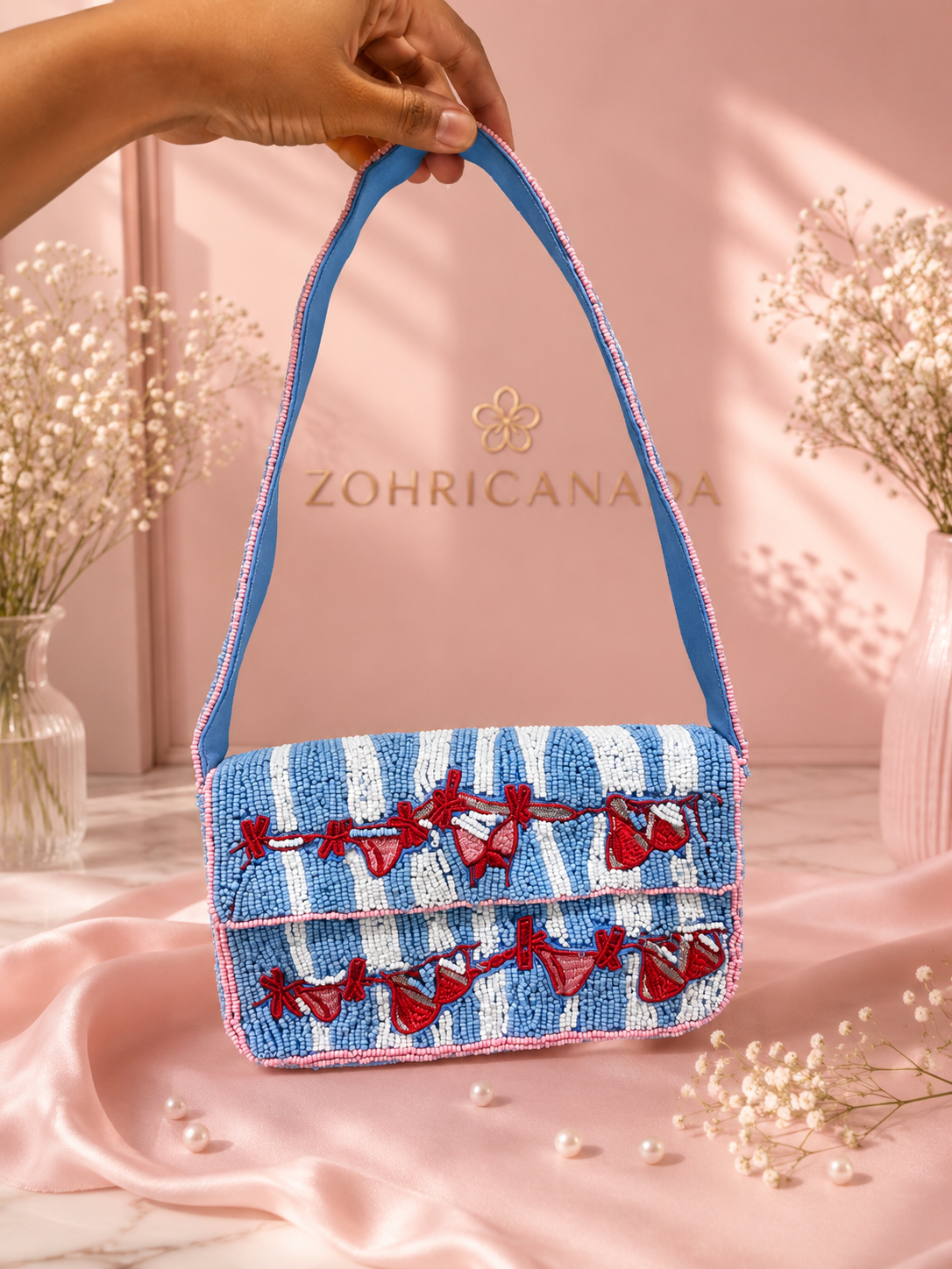 Handmade Blue & White Striped Beaded Shoulder Bag for Women | Beach & Resort Handbag — A hand holds a small blue and white striped beaded handbag with red bow embellishments and a blue shoulder strap, set against a pink backdrop with the brand name ZOHRICANADA visible. — Zohricanada